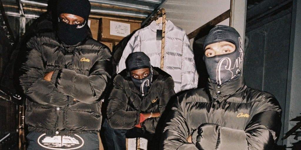 Three men wearing black Corteiz puffer jackets and branded balaclavas pose with arms crossed in an urban warehouse setting.