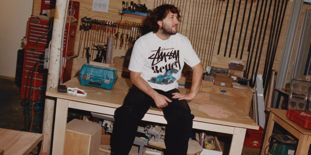 Man wearing a Stüssy graphic T-shirt sitting on a workbench in a workshop.